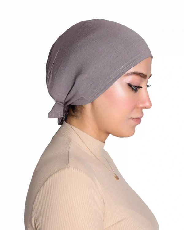 A woman models a Chic Grey Turban Cap for Women, shown in a profile view with a hand near her chin, emphasizing the tidy tie-back detail and smooth silhouette.
